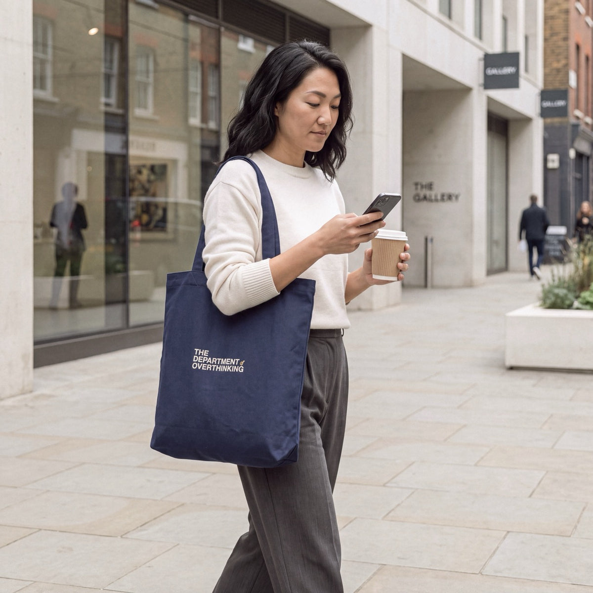 Woman carrying a navy embroidered Department of Overthinking tote bag while checking her phone