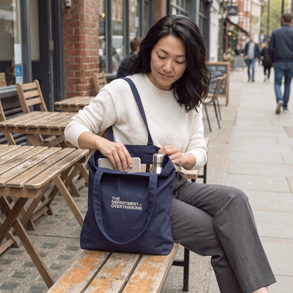 Woman reaching into a navy Department of Overthinking tote bag while sitting outdoors