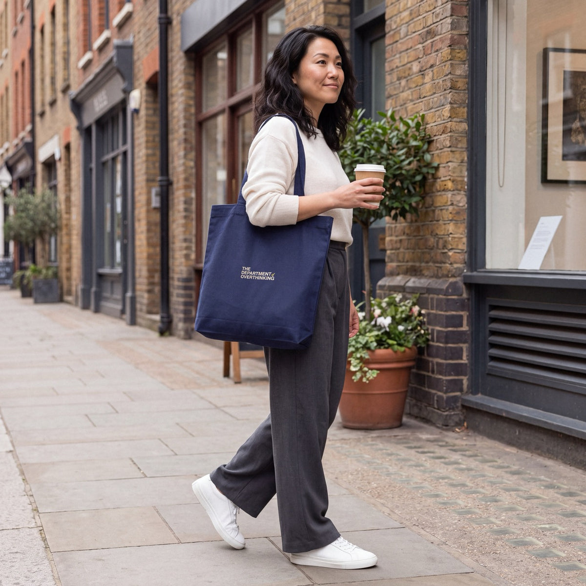 Woman carrying a navy Department of Overthinking embroidered tote bag while walking with a coffee