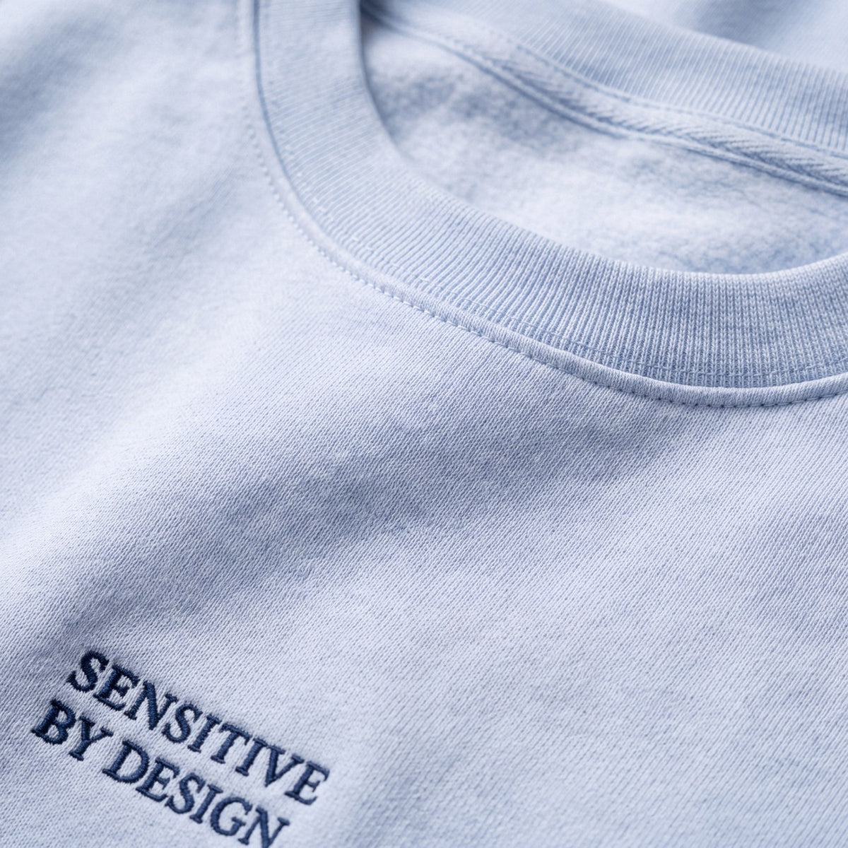 Close-up of SENSITIVE BY DESIGN. embroidery on light blue sweatshirt