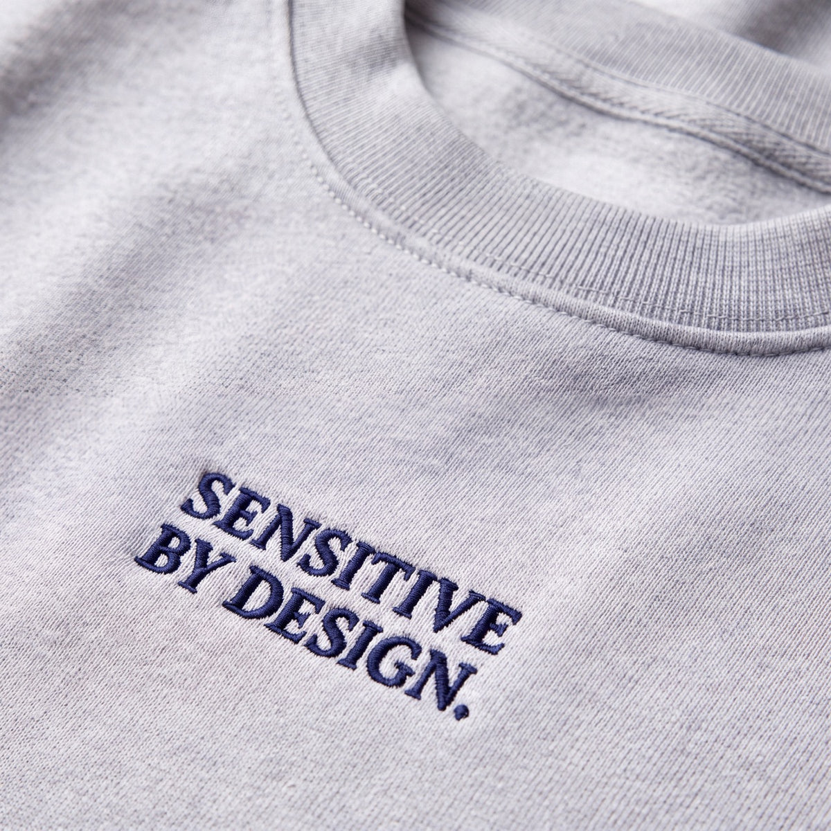 Close-up of SENSITIVE BY DESIGN. embroidery on ash sweatshirt