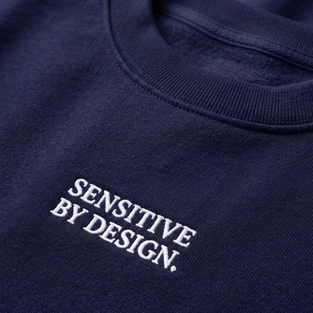 Close-up of SENSITIVE BY DESIGN. embroidery on navy sweatshirt