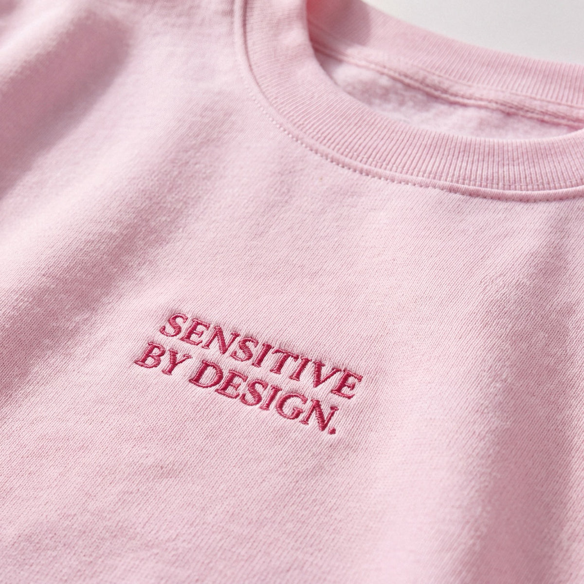 Close-up of SENSITIVE BY DESIGN. embroidery on light pink sweatshirt